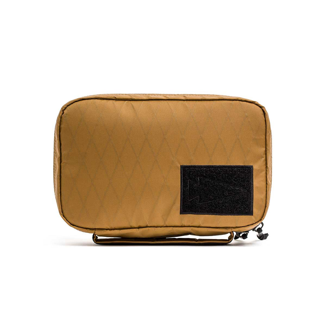 The GORUCK GR1 Field Pocket - X-PAC is a tan rectangular pouch with a black front patch, made from waterproof fabric and featuring a bottom right zipper—perfect as an electronics organizer.