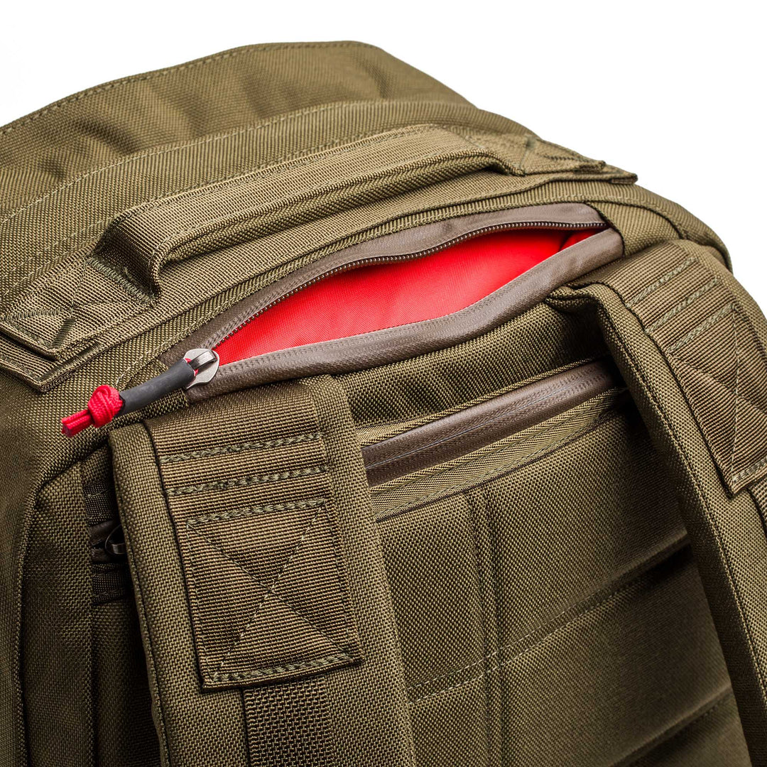 Close-up of the GORUCK GR2 travel backpack in olive green, showing its red interior through a partially open zipper pocket.