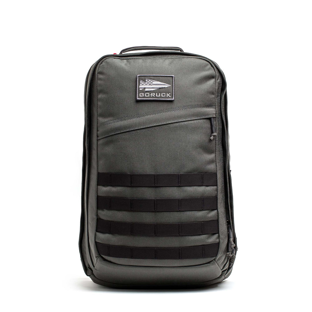 A GORUCK GR2 backpack in gray with black webbing and a front zip pocket stands upright on a white background.