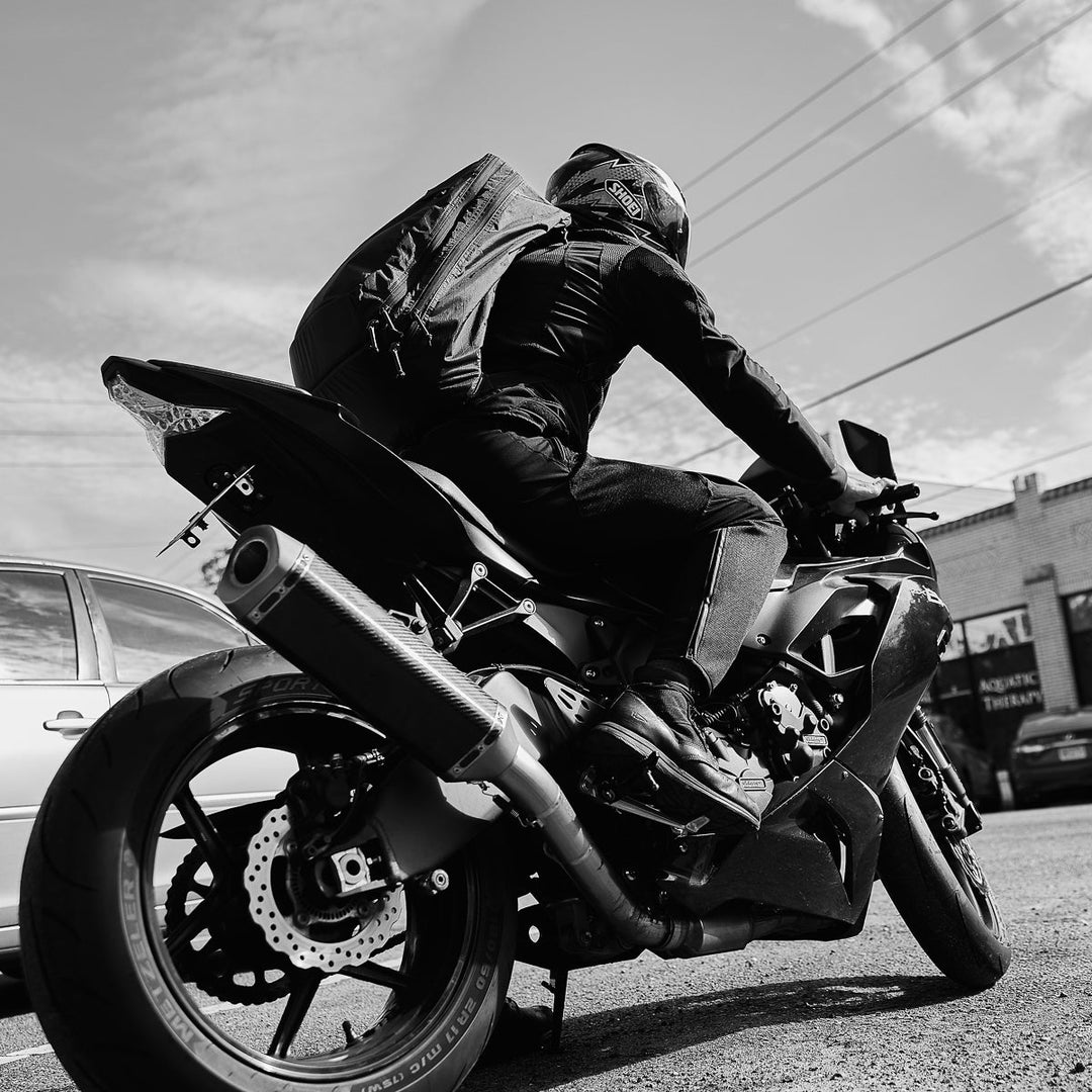 A motorcyclist rides a sport bike on a city street, viewed from a low rear angle, carrying the GORUCK GR2 X-PAC ruck made with waterproof X-PAC material.