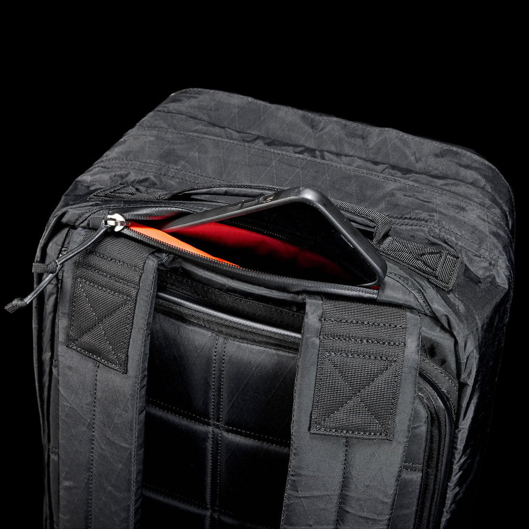 The GORUCK GR2 X-PAC, a black waterproof backpack, features a zippered pocket partially open to reveal a tablet with a red case inside—ensuring your gear stays safe and stylish wherever you go.