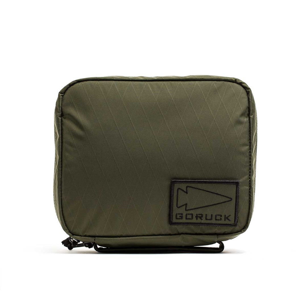 The GORUCK GR2 Field Pocket - X-PAC is an olive green square pouch with a GORUCK patch and arrowhead logo, made from waterproof fabric—ideal for organizing electronics or carrying camera gear.