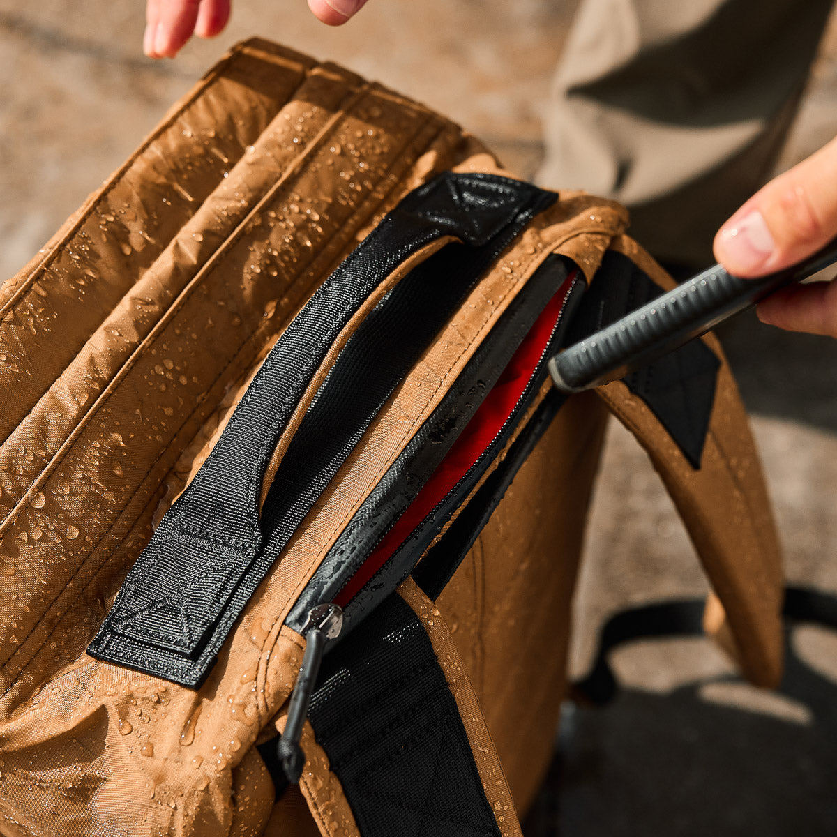 A person opens a wet tan backpack, revealing a red folder inside.