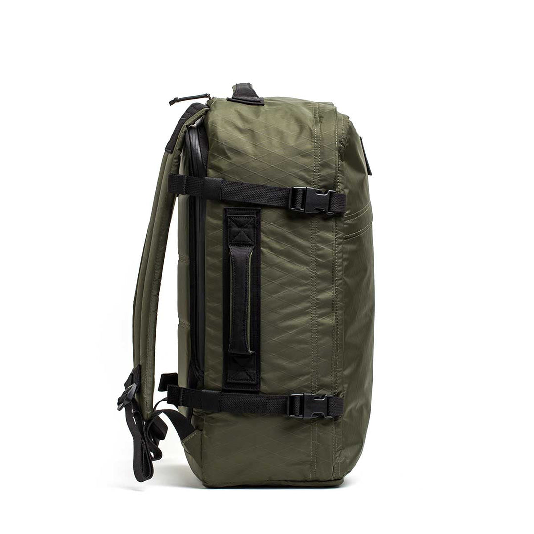 The GR3 - X-PAC olive green carry-on backpack stands upright in a side view, showing black straps and a handle; it includes a convenient laptop compartment.