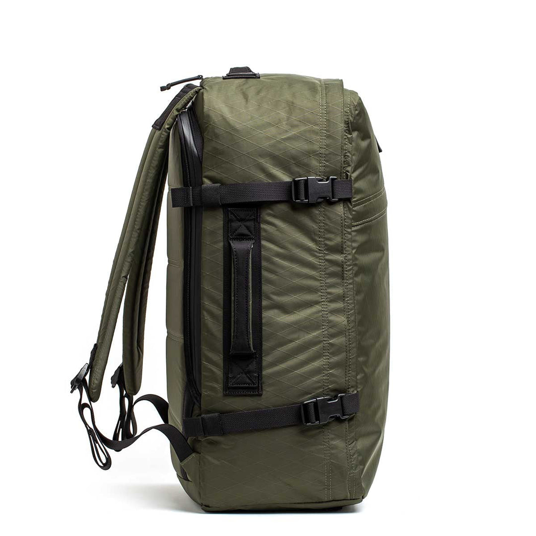 The GR3 - X-PAC carry-on backpack in olive green is shown from the side, featuring black straps, a top handle, and a waterproof construction.