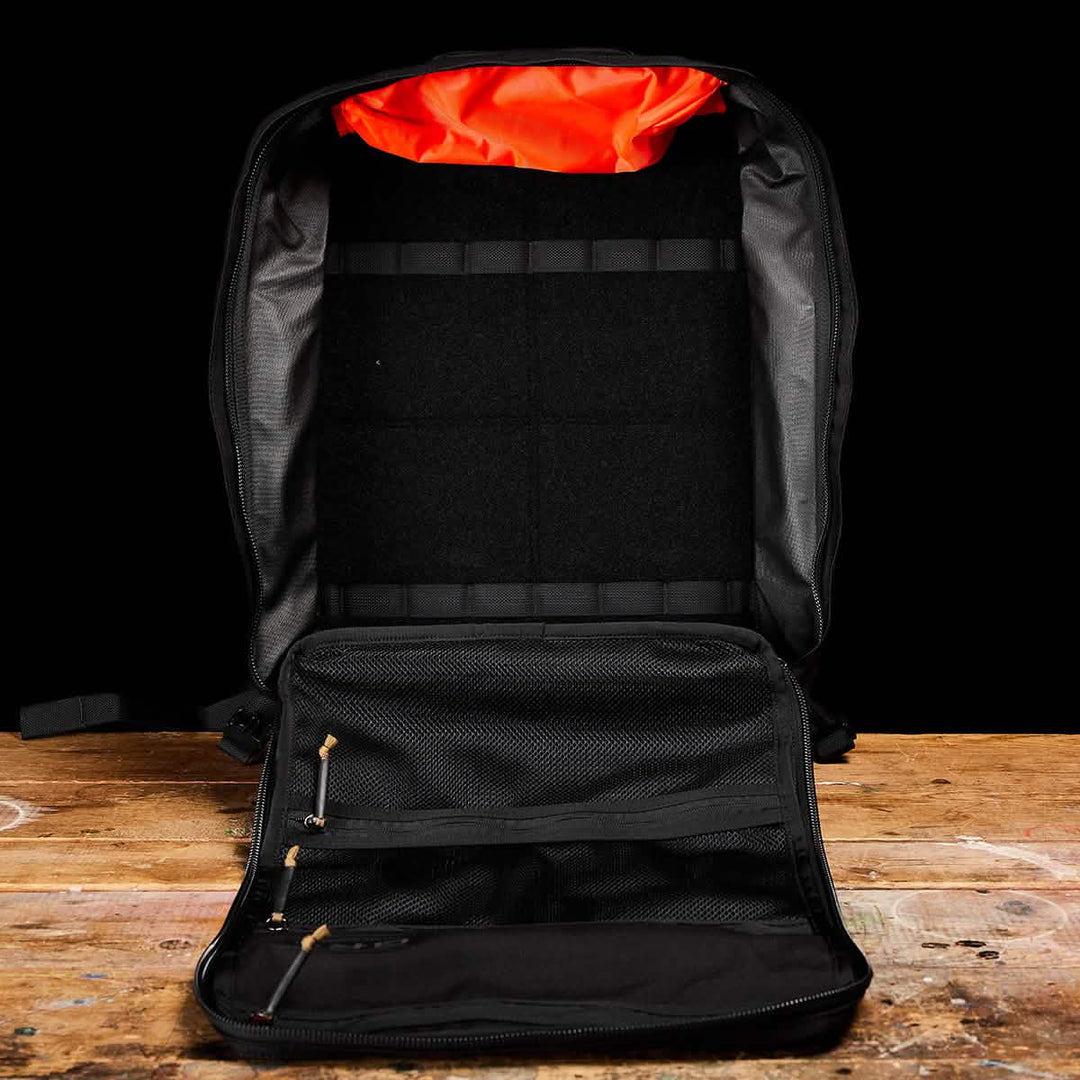 A GORUCK GR3 - Cordura open black travel backpack with orange lining and waterproof zippers sits on a wooden surface against a black background.