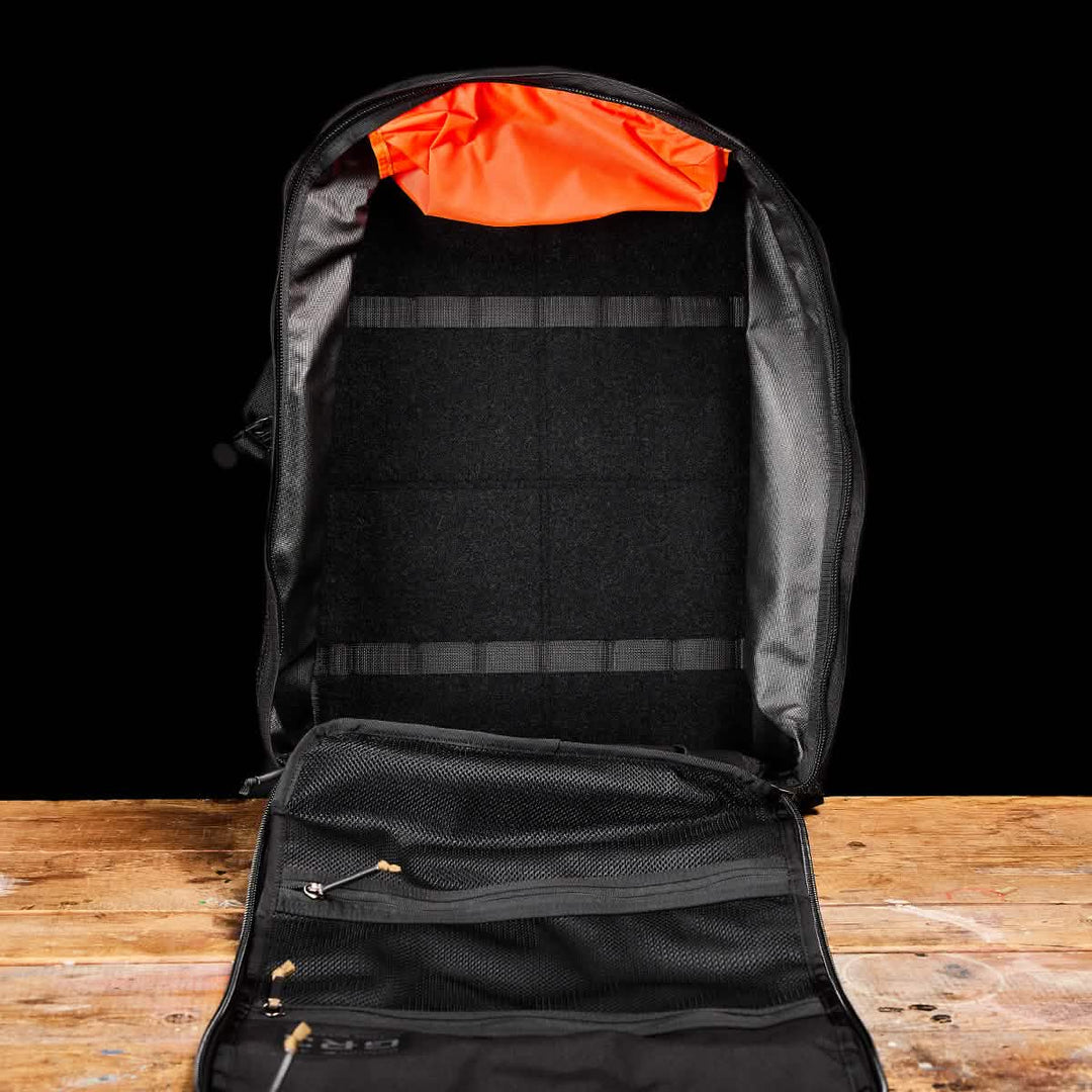 The GORUCK GR3 - Cordura carry-on rucksack in black, with an orange inner lining, is shown open on a wooden surface, highlighting its interior pockets and waterproof zippers.