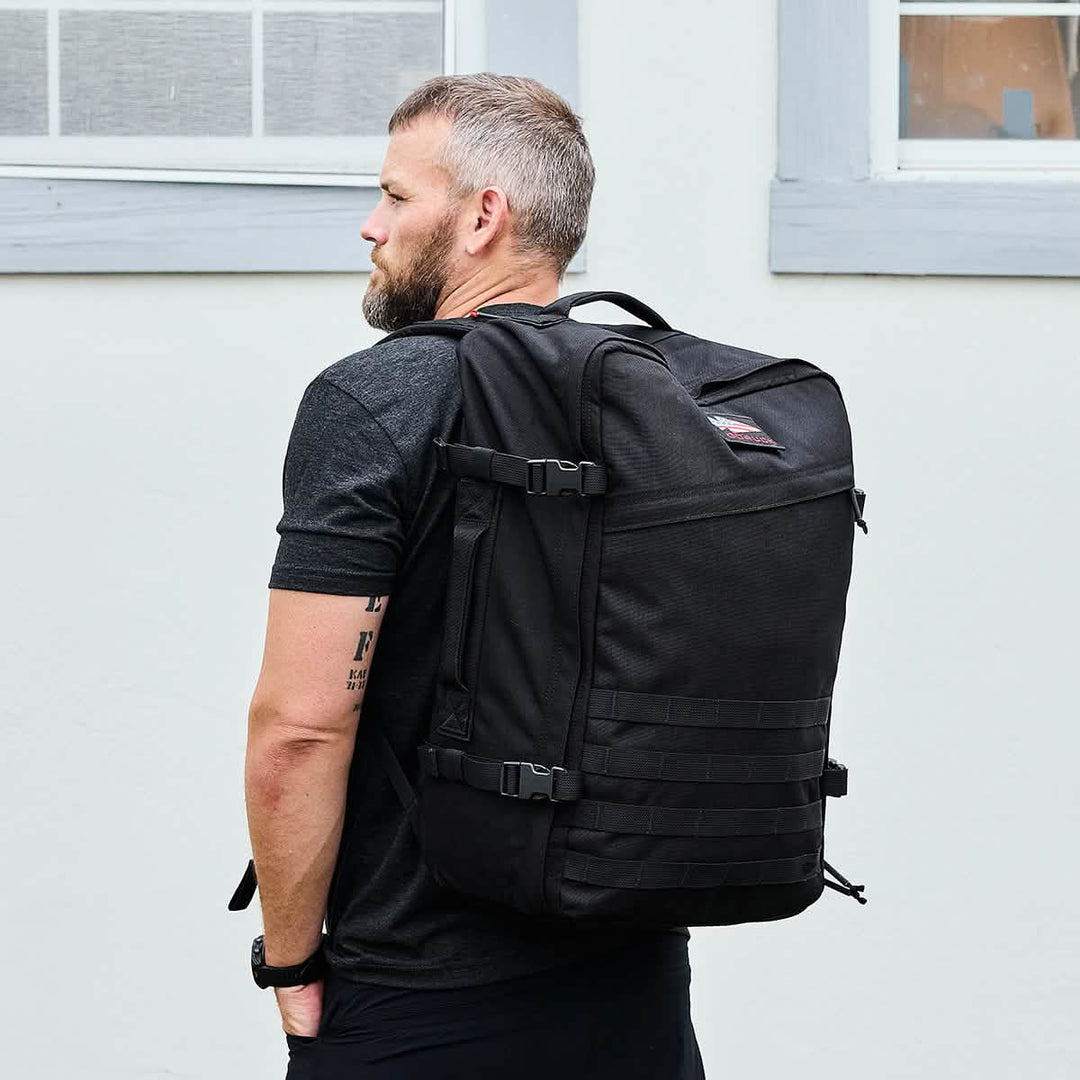 A bearded man wearing a black t-shirt and carrying the GORUCK GR3 - Cordura rucksack with waterproof zippers stands in front of a light-colored wall.