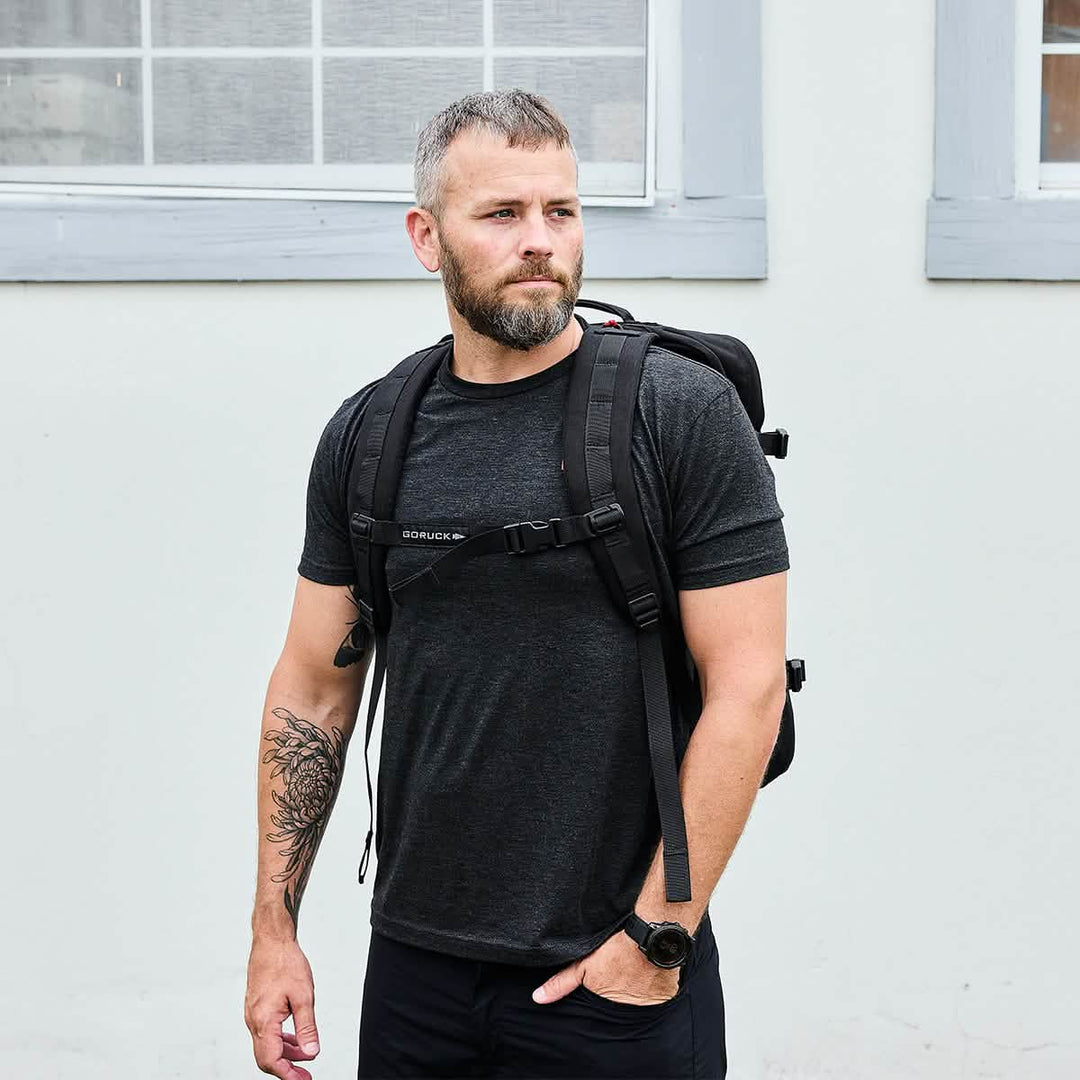 A bearded man in a black t-shirt carries the GORUCK GR3 - Cordura travel backpack with waterproof zippers while standing outside by a white building with windows.