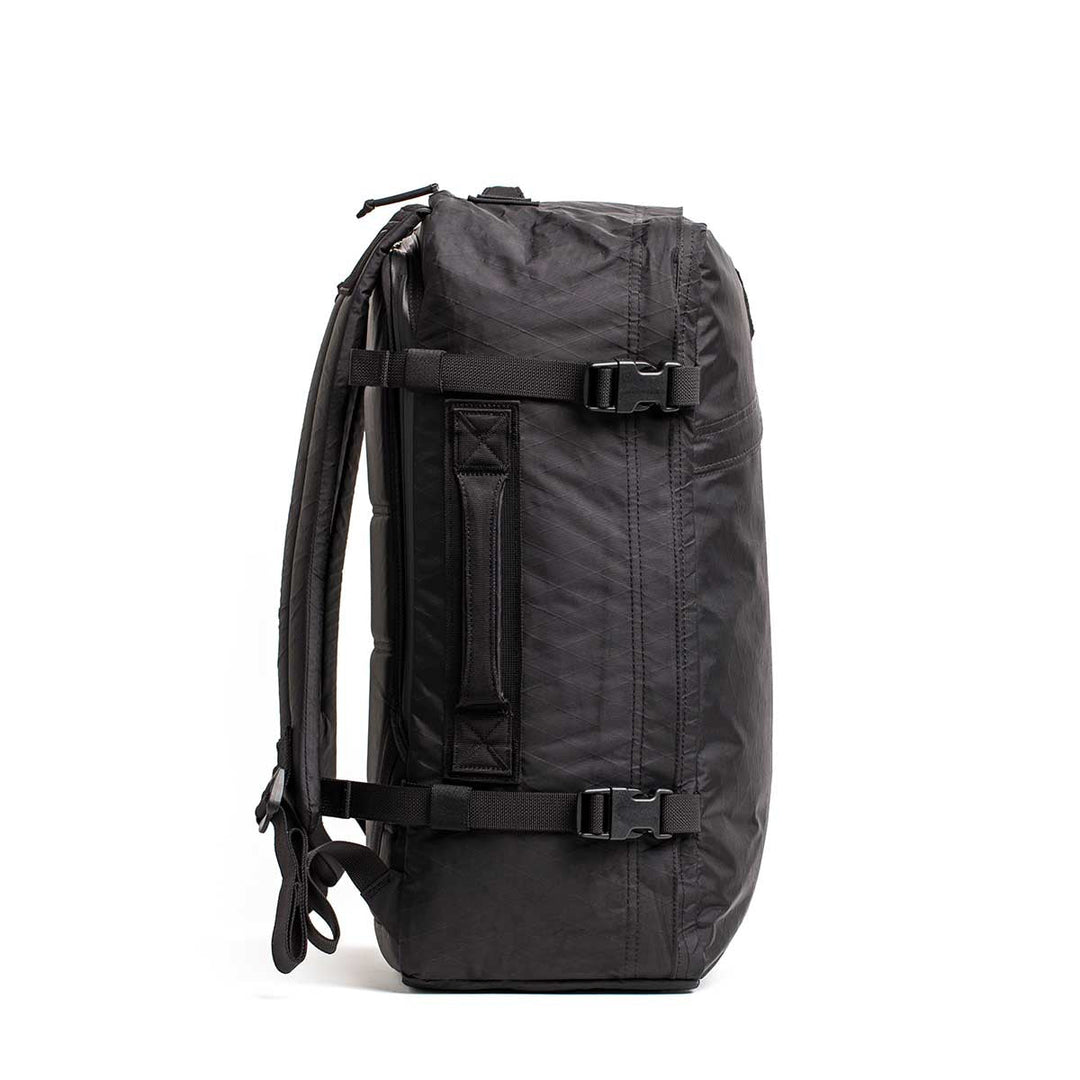 The GORUCK GR3 - X-PAC carry-on backpack is shown from the side on a white background, highlighting buckles, handle, and shoulder straps. Made with ultra-waterproof material, it comes with a lifetime guarantee for durability.