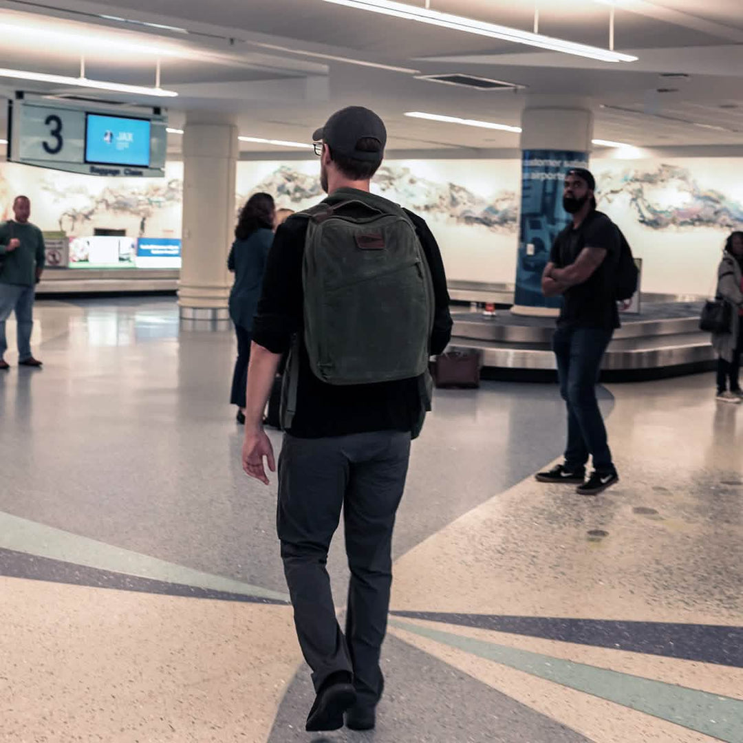 A person with a green GR1 USA - Heritage by GORUCK casually strolls through the airport baggage claim area, surrounded by a few other travelers.