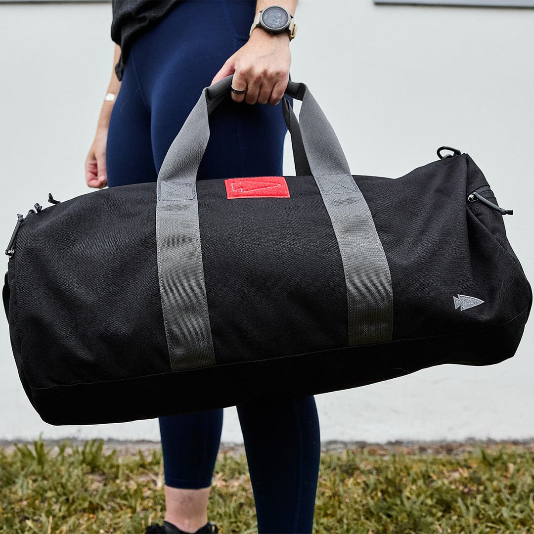 A person is outdoors holding a sleek black GORUCK Gym Bag - Cordura with gray straps, while sporting a watch and blue leggings.