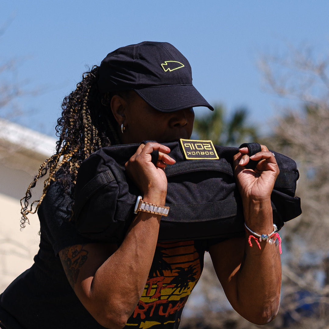 Wearing the GORUCK Performance Running Hat - ToughMesh, a person lifts a weighted sandbag outdoors on a sunny day.