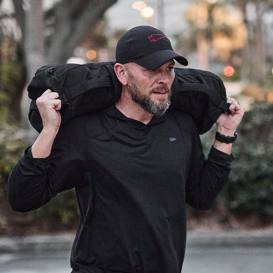 A man wearing the GORUCK Performance Running Hat - ToughMesh carries a heavy sandbag on his shoulders outdoors at dusk.