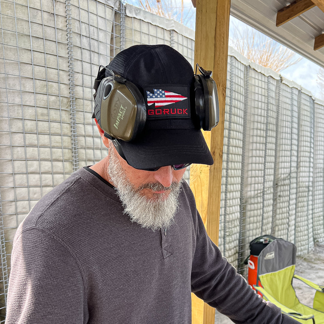 A man with a gray beard, sunglasses, and earmuffs on his cap stands outdoors by a wire fence, wearing the GORUCK Cadre Hat made from durable 500D CORDURA®.