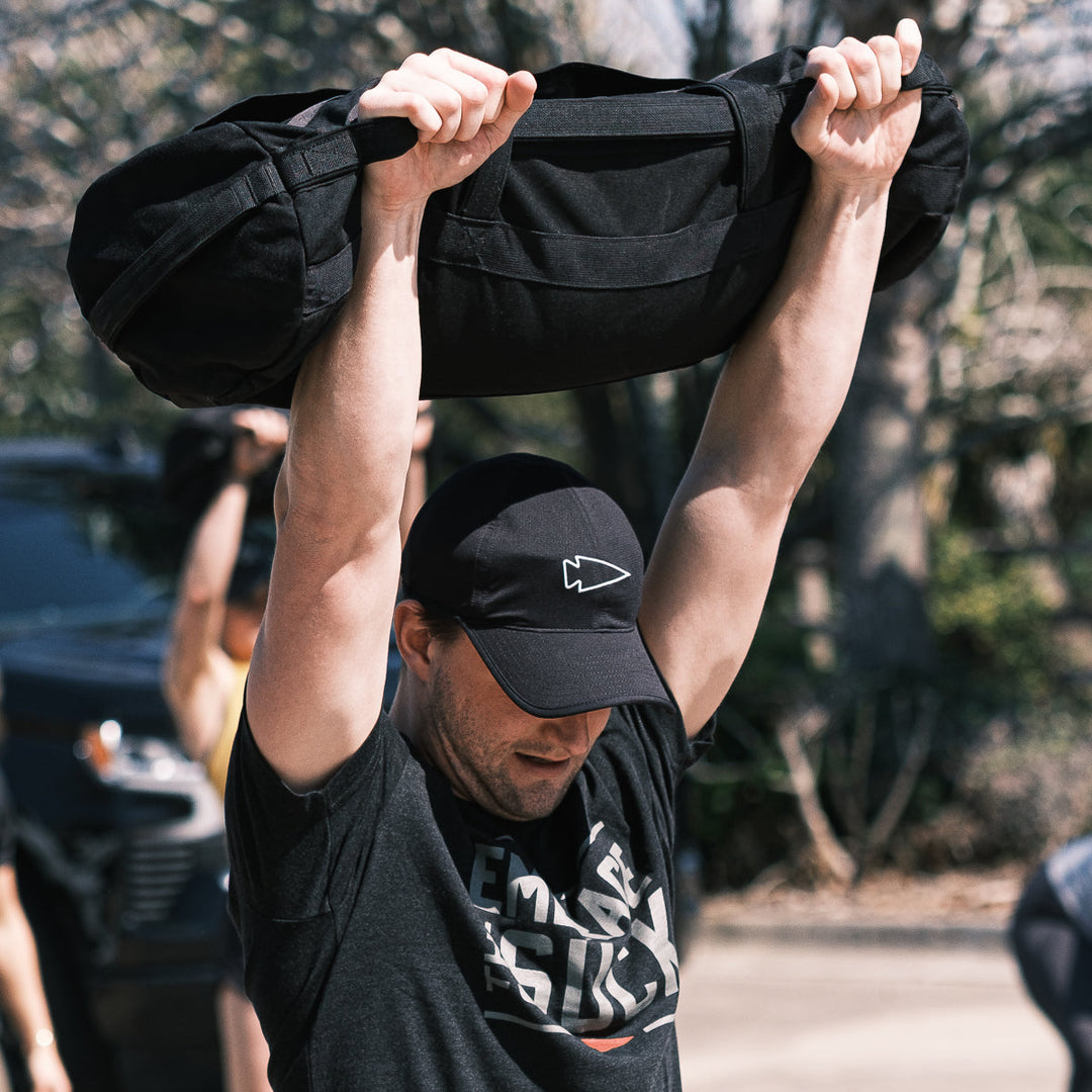 Man lifting a heavy sandbag overhead outdoors, wearing a black t-shirt and the GORUCK Performance Running Hat - ToughMesh for comfort during intense workouts.