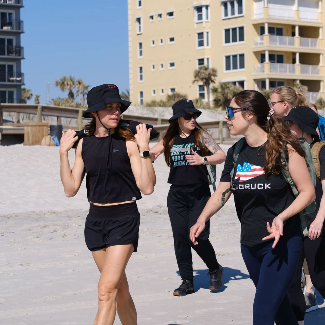 Four women in athletic wear, including one wearing the GORUCK Boonie Hat - Slick - ToughDry with 50+ UPF protection, walk and chat on a sunny beach with buildings in the background.