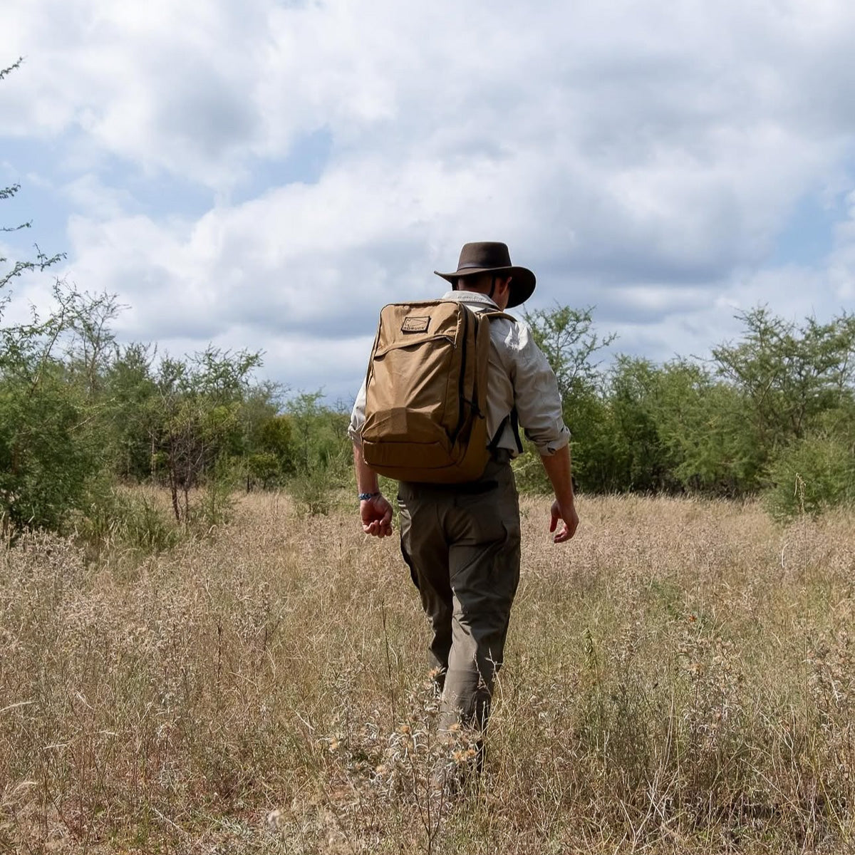 Man rucking outdoors with tan GORUCK backpack, wearing hat, khaki shirt, and rugged pants
