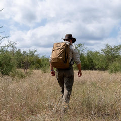 Man rucking outdoors with tan GORUCK backpack, wearing hat, khaki shirt, and rugged pants
