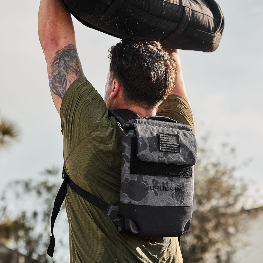 A man lifts a heavy sandbag overhead outdoors, wearing a green shirt and the GORUCK Ruck Plate Carrier 3.0—a small black backpack with padded shoulder straps for comfort.