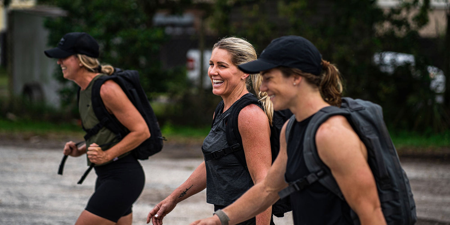 Three women wearing athletic gear and backpacks walk outdoors, smiling and enjoying themselves.