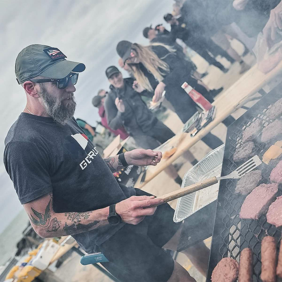 Man in GORUCK shirt and cap grilling burgers at a beach community event
