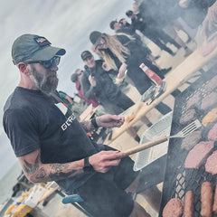 Man in GORUCK shirt and cap grilling burgers at a beach community event