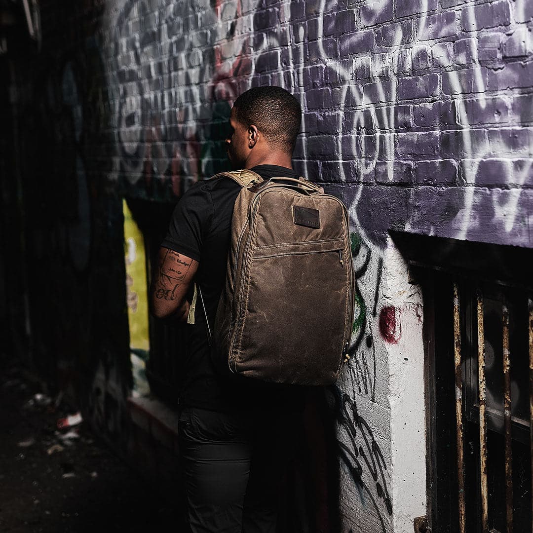 An individual carrying a GORUCK GR1 USA - Heritage rucksack stands in an alley adorned with graffiti-covered walls.