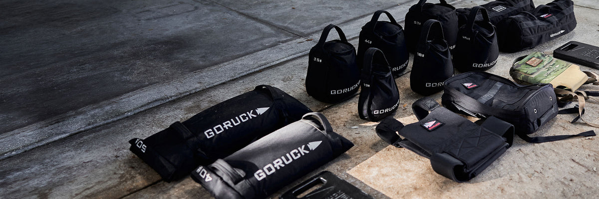Several black GORUCK sandbags and weighted vests are arranged on a concrete floor in natural light.
