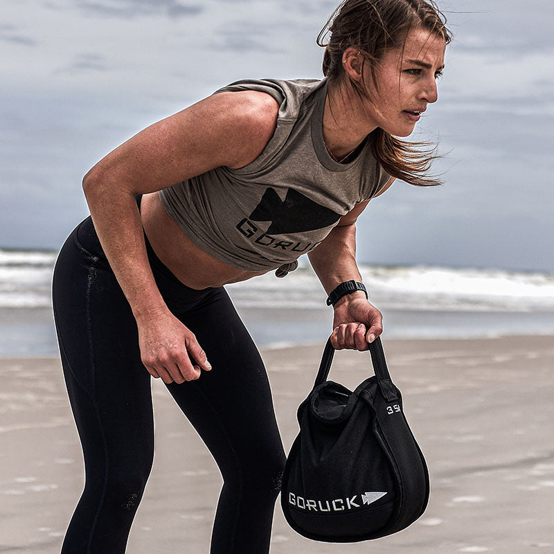Woman exercising on a beach, holding a black GORUCK sandbag, wearing athletic clothing.