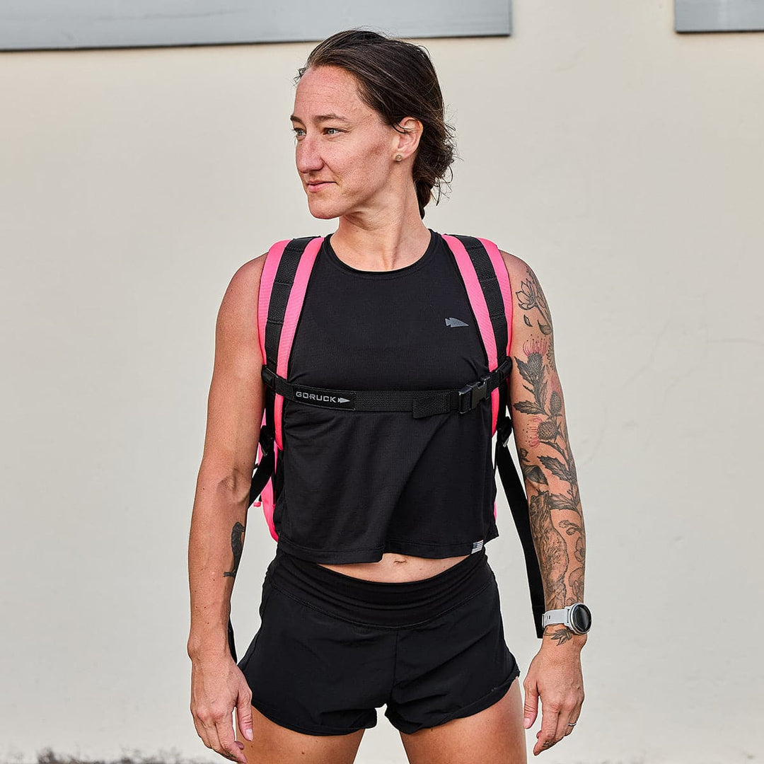 A person with tattoos stands outdoors against a plain off-white wall, wearing a black sleeveless crop top and shorts, and carrying the GR0 - Cordura compact backpack in bright pink by GORUCK. They have short hair and a neutral expression.
