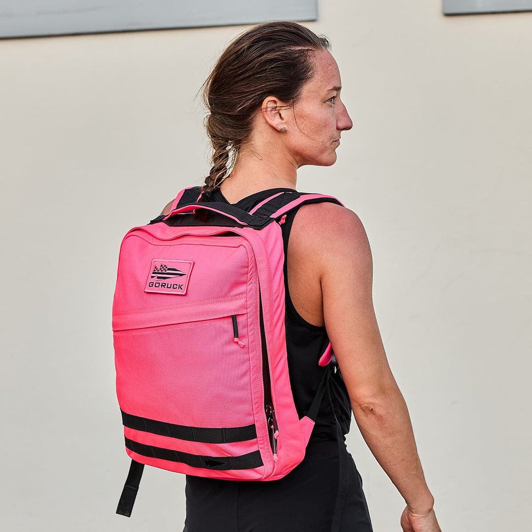 A person with braided hair is wearing a bright pink GR0 - Cordura backpack by GORUCK, known for its bombproof laptop compartment, along with a black sleeveless top. They stand against a light gray wall, looking to the side. The backpack includes black straps and logos, making it perfect for minimalist EDC enthusiasts.