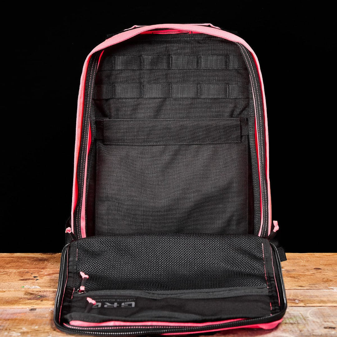 Displayed upright on a wooden surface against a black background is the GR0 - Cordura by GORUCK, an open pink and black compact backpack featuring multiple compartments, mesh pockets, and a bombproof laptop compartment.