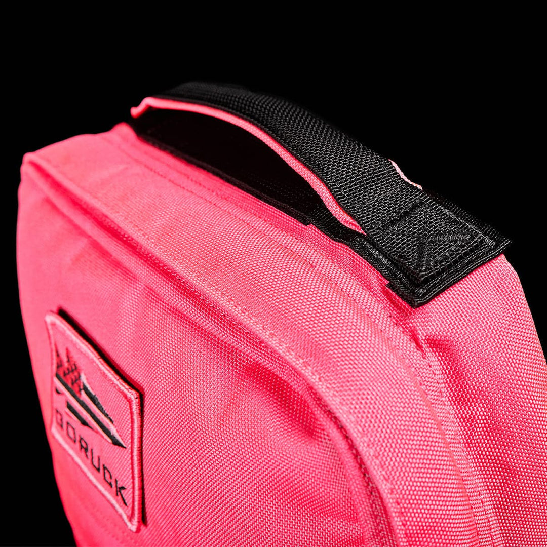 Close-up of the GR0 - Cordura backpack by GORUCK in pink against a black background. The black handle and a mountain design patch are highlighted. The fabric looks durable, featuring prominent stitching and an ultra-protective laptop compartment, ideal for minimalist EDC enthusiasts.