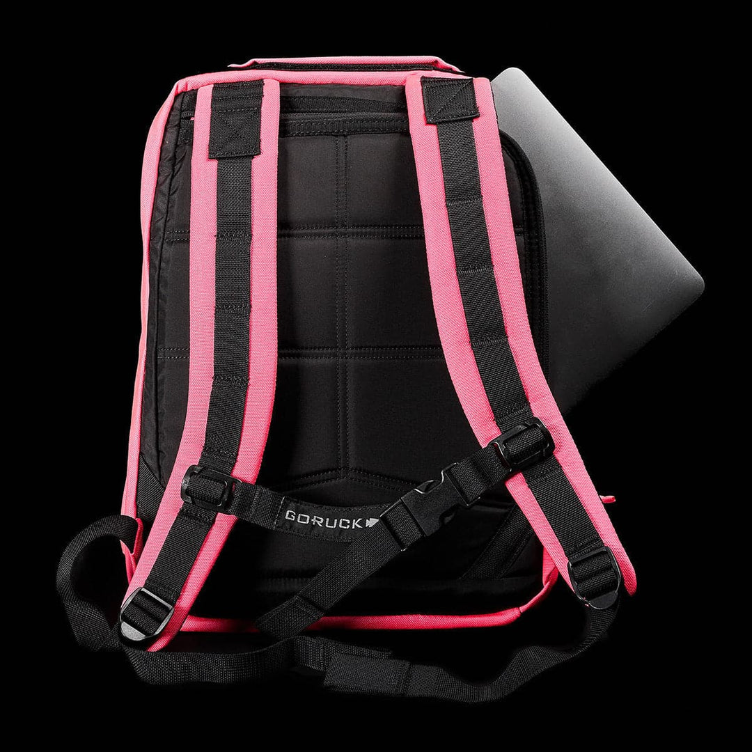 The GR0 - Cordura by GORUCK, a black, compact backpack featuring a bombproof laptop compartment, is displayed against a black background. The minimalist EDC appeal is enhanced by bright pink padded straps and a silver laptop partially tucked into the main compartment.