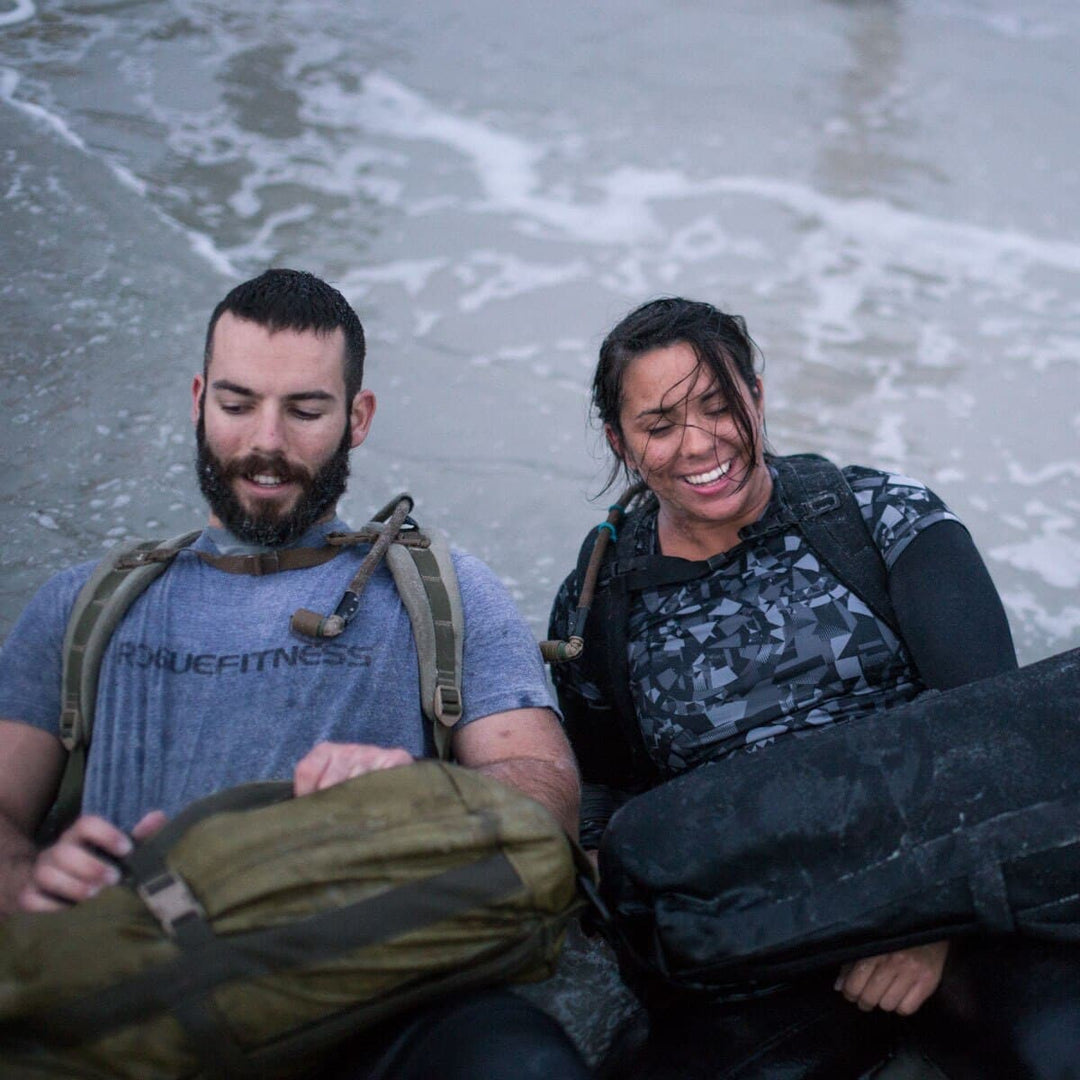 Two people in wet clothing are sitting on a beach, their backpacks hinting at an adventurous journey. Despite the rain, they're smiling, perhaps relieved to have a GORUCK hydration valve mouthpiece tucked away with a replacement ready for whatever comes next.
