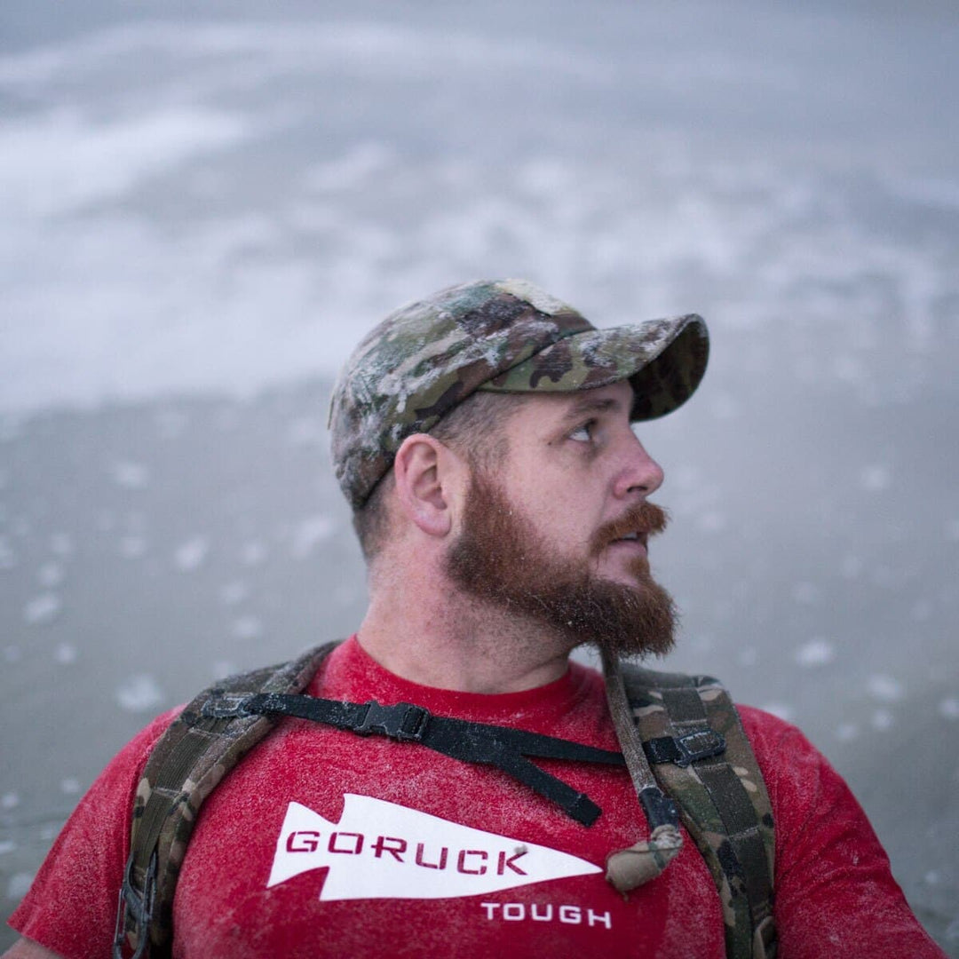 A bearded man in a camouflage cap and a red GORUCK shirt is on the beach, subtly adjusting his GORUCK Hydration Valve Mouthpiece while looking to the side.