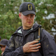 A man in a cap, wearing a dark jacket, examines a small GORUCK Hydration Valve Mouthpiece attached to his shoulder while outdoors in a forested area.