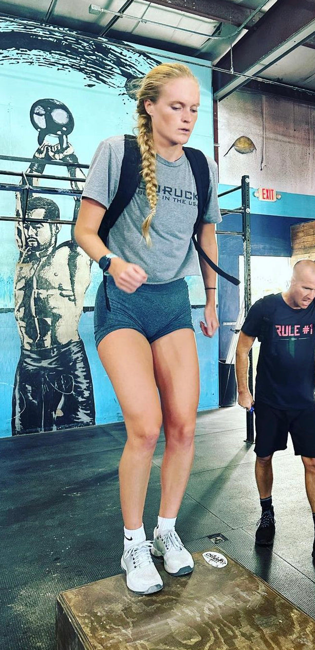A woman with a long braid steps up on a box in a gym, wearing workout clothes and a weighted vest.