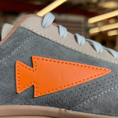 Close-up of the Mackall 45 Fog sneaker featuring gray suede, white laces, and an orange arrow-shaped patch on the side.