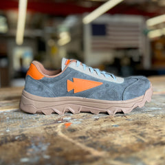 The Mackall 45 Fog features gray suede with orange accents and a thick tan sole, displayed on a worn wooden surface.
