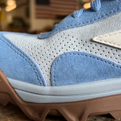 Close-up of the Mackall 45 Americana sneaker in blue and gray with a brown sole, displayed indoors against a blurred American flag backdrop.