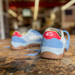 Two Mackall 45 Americana sneakers in blue and white are displayed on a wooden table, featuring a red “45” on the heel of the right shoe.