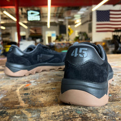 Mackall 45 Black + Gum sneakers with “415” on the heel rest on a worn table; an American flag hangs in the background.