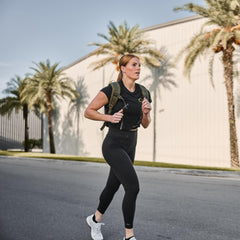 A woman jogs down a palm-lined street wearing squat-proof Training Leggings and carrying a backpack, ready for any workout.