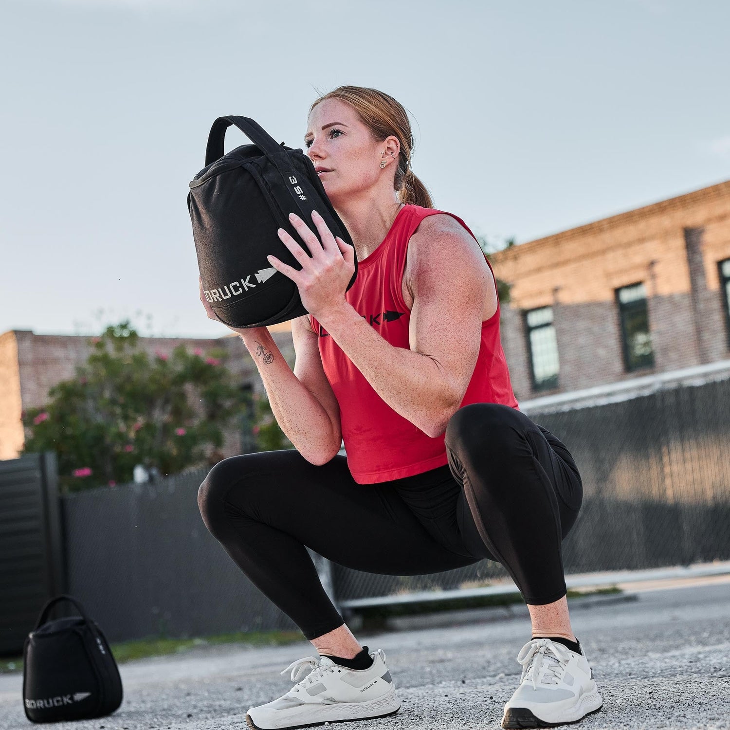 A woman in athletic wear squats outdoors, holding a black weighted bag to her chest and showing off her squat-proof Training Leggings, designed for even the toughest workouts.