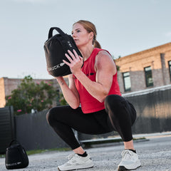 A woman in athletic wear squats outdoors, holding a black weighted bag to her chest and showing off her squat-proof Training Leggings, designed for even the toughest workouts.