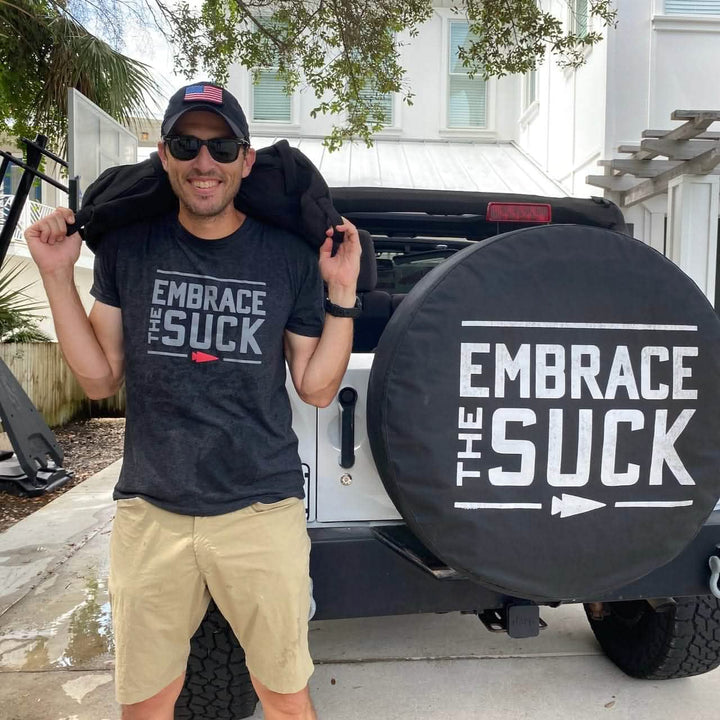 A man in sunglasses and an Embrace the Suck Tee - Tri-Blend by vendor-unknown stands by a jeep with "Embrace the Suck" on his shirt and tire cover, showcasing the USMC motto.