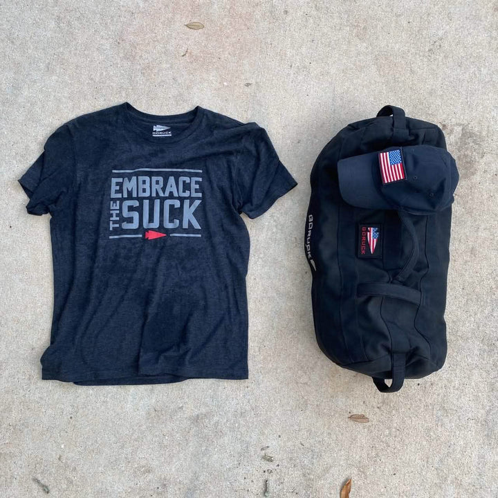The Embrace the Suck Tee - Tri-Blend by vendor-unknown, a black duffel bag, and an American flag patch cap.