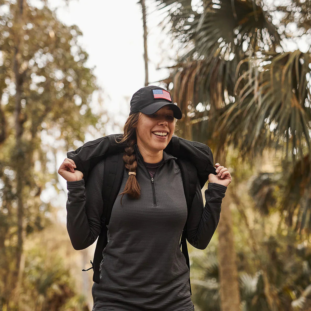 Woman rucking outdoors with GORUCK gear, wearing black cap and backpack on forest trail